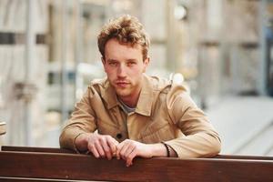 Elegant young man in formal classy clothes leaning on wooden fence outdoors in the city photo