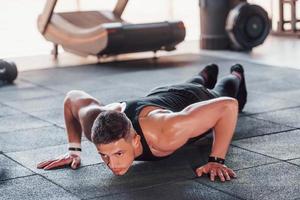Doing push-ups. Young sportive strong man in black wear have workout day in gym photo