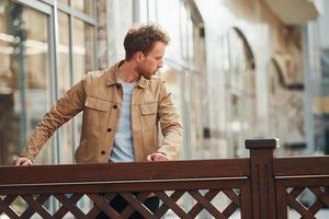 Elegant young man in formal classy clothes leaning on wooden fence outdoors in the city photo