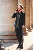 Elegant young man in formal classy clothes standing and talking by the phone outdoors in the city photo