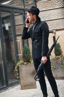 Talking by phone. Elegant young man in black hat and clothes with umbrella in hands outdoors in the city photo