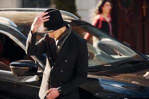 parado cerca del auto negro. joven elegante con ropa elegante y formal al aire libre en la ciudad foto