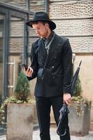 Talking by phone. Elegant young man in black hat and clothes with umbrella in hands outdoors in the city photo