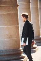 con la computadora portátil en las manos. joven elegante con gafas y ropa formal elegante al aire libre en la ciudad foto