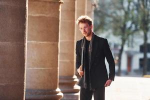 With laptop in hands. Elegant young man in glasses adn in formal classy clothes outdoors in the city photo