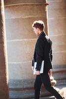 With laptop in hands. Elegant young man in glasses adn in formal classy clothes outdoors in the city photo