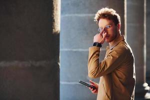 Nice sunshine. Holding phone in hand. Elegant young man in formal classy clothes outdoors in the city photo