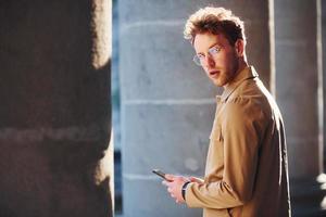 Nice sunshine. Holding phone in hand. Elegant young man in formal classy clothes outdoors in the city photo