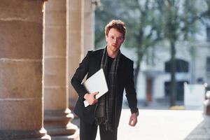 With laptop in hands. Elegant young man in glasses adn in formal classy clothes outdoors in the city photo