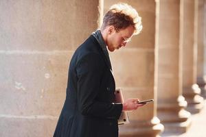 With laptop and phone in hands. Elegant young man in glasses adn in formal classy clothes outdoors in the city photo