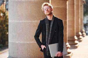 With laptop in hands. Elegant young man in glasses adn in formal classy clothes outdoors in the city photo