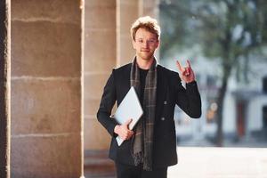 con la computadora portátil en las manos. un joven amable y elegante con gafas y ropa elegante y formal al aire libre en la ciudad foto