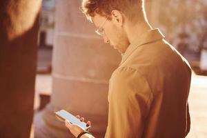 Nice sunshine. Holding phone in hand. Elegant young man in formal classy clothes outdoors in the city photo