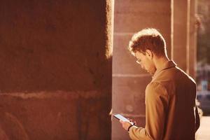 Nice sunshine. Holding phone in hand. Elegant young man in formal classy clothes outdoors in the city photo