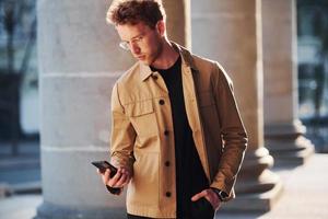 Nice sunshine. Holding phone in hand. Elegant young man in formal classy clothes outdoors in the city photo