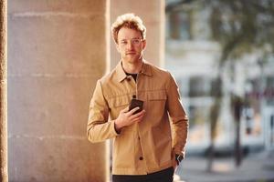 Nice sunshine. Holding phone in hand. Elegant young man in formal classy clothes outdoors in the city photo