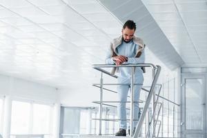 Leaning on silver colored railings. Young handsome man in formal clothes indoors in the office at daytime photo