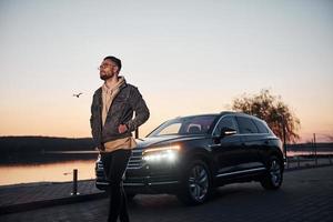 Handsome unshaved man in fashionable clothes walks near his black car at evening time photo