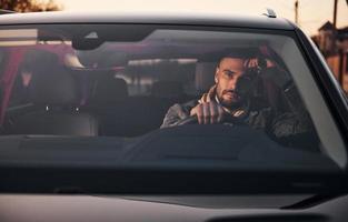 Front view of handsome unshaved man in fashionable clothes that riding his automobile photo