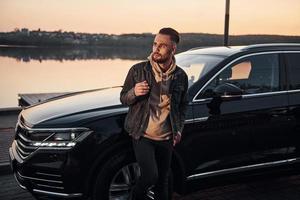 Handsome unshaved man in fashionable clothes standing near his black car and smoking photo