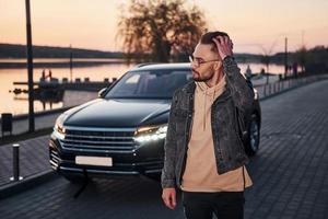 Handsome unshaved man in fashionable clothes walks near his black car at evening time photo