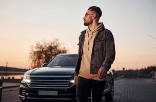 Handsome unshaved man in fashionable clothes walks near his black car at evening time photo