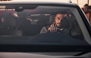 Front view of handsome unshaved man in fashionable clothes that riding his automobile photo