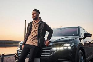 Handsome unshaved man in fashionable clothesleaning on his his black car photo