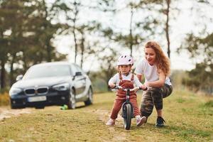 coche en el fondo. madre con camisa blanca enseñando a su hija a andar en bicicleta al aire libre foto