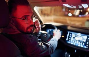 Talking by phone. Red lighting. Handsome unshaved man in fashionable clothes riding his automobile photo