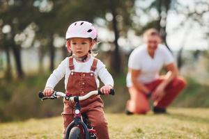 padre con camisa blanca enseñando a su hija a andar en bicicleta al aire libre foto