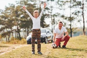 Playing soccer. Father with his son spending weekend together outdoors near forest at daytime photo