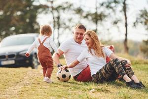 jugando con balón de fútbol. padre y madre pasan el fin de semana al aire libre cerca del bosque con su hija foto