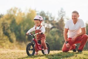 Beautiful sunlight. Father in white shirt teaching daughter how to ride bicycle outdoors photo