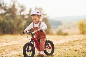 Happy little girl in protective hat riding her bike outdoors at sunny day near forest photo