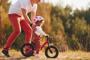 Beautiful sunlight. Father in white shirt teaching daughter how to ride bicycle outdoors photo