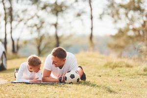 With soccer ball. Father with his son lying down on the ground together outdoors near forest at daytime photo