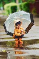 Kid in yellow waterproof cloak, boots and with umbrella playing outdoors after the rain photo