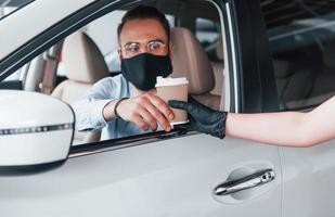 Buying fresh drink. Young handsome man in formal clothes and protective mask sitting in brand new automobile photo