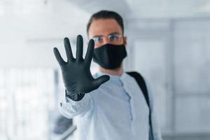 Showing stop gesture by hand. In protective mask and gloves. Young handsome man in formal clothes indoors in the office at daytime photo