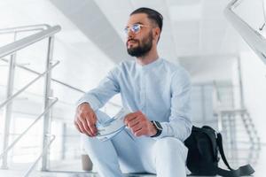 Holding plane tickets in hand. Sitting on stairs and waiting. Young passenger in formal clothes indoors in the office at daytime photo