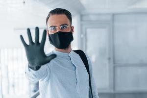 Showing stop gesture by hand. In protective mask and gloves. Young handsome man in formal clothes indoors in the office at daytime photo