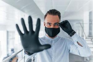 Showing stop gesture by hand. In protective mask and gloves. Young handsome man in formal clothes indoors in the office at daytime photo