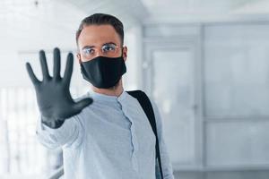 Showing stop gesture by hand. In protective mask and gloves. Young handsome man in formal clothes indoors in the office at daytime photo