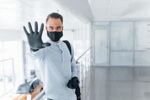 Showing stop gesture by hand. In protective mask and gloves. Young handsome man in formal clothes indoors in the office at daytime photo