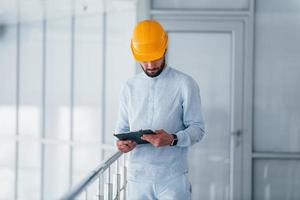Holding notepad. Engineer in white clothes and orange protective hard hat standing and working indoors photo