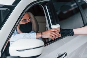 Buying fresh drink. Young handsome man in formal clothes and protective mask sitting in brand new automobile photo