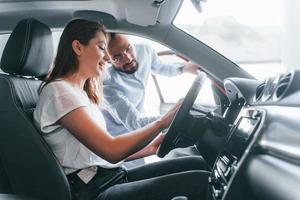 vista lateral de una joven positiva que tiene una visita en autosalon y elige auto. gerente ayudándola foto