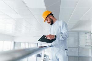 Holding notepad. Engineer in white clothes and orange protective hard hat standing and working indoors photo