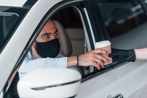 Buying fresh drink. Young handsome man in formal clothes and protective mask sitting in brand new automobile photo